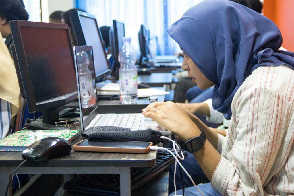 A young woman in hijab using a laptop in a computer classroom setting.