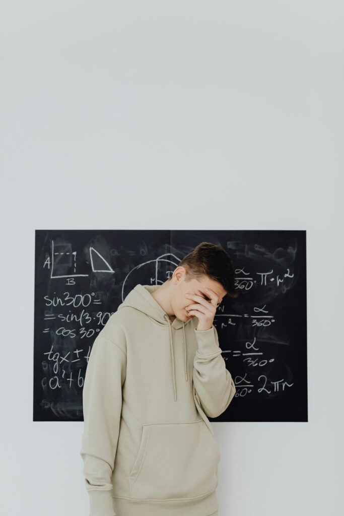 A young boy in a hoodie stands covering his face in frustration before a math equation-filled chalkboard.