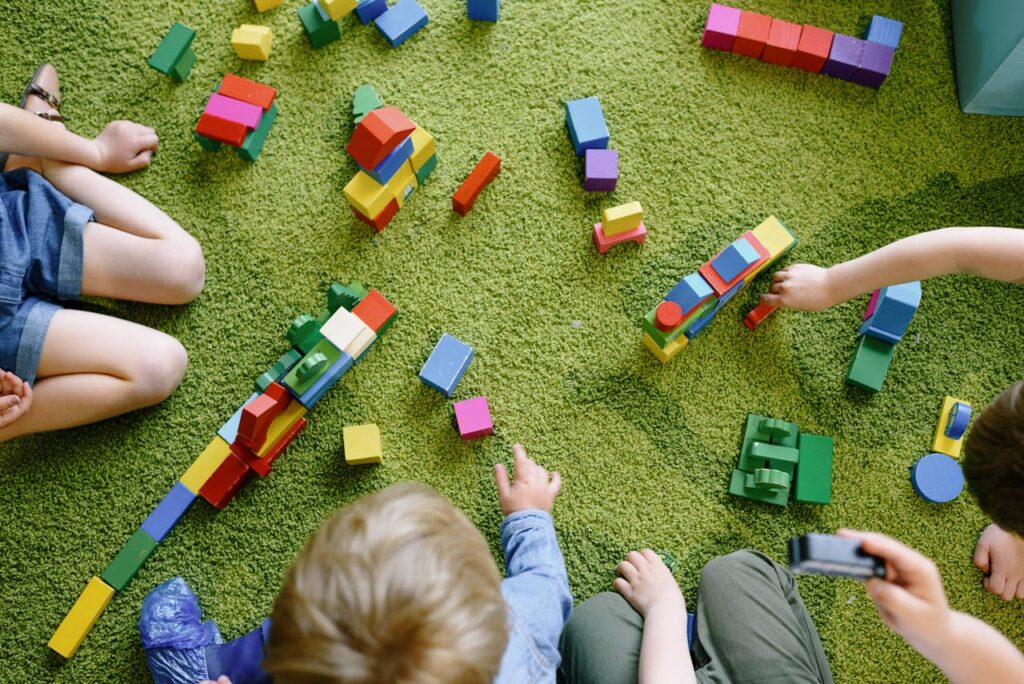 Children playing with wooden blocks on a green carpet in a classroom setting.